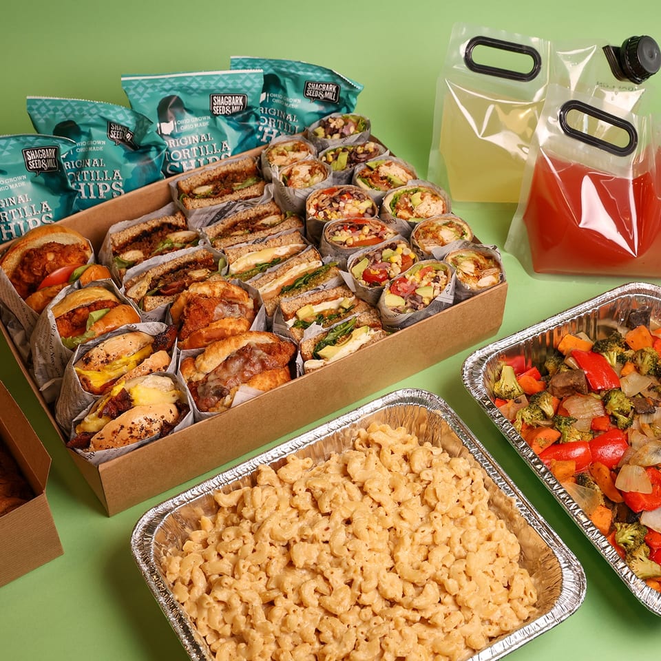 Catering spread featuring sandwiches, wraps, tortilla chips, macaroni and cheese, roasted vegetables, cookies, and bottled drinks.