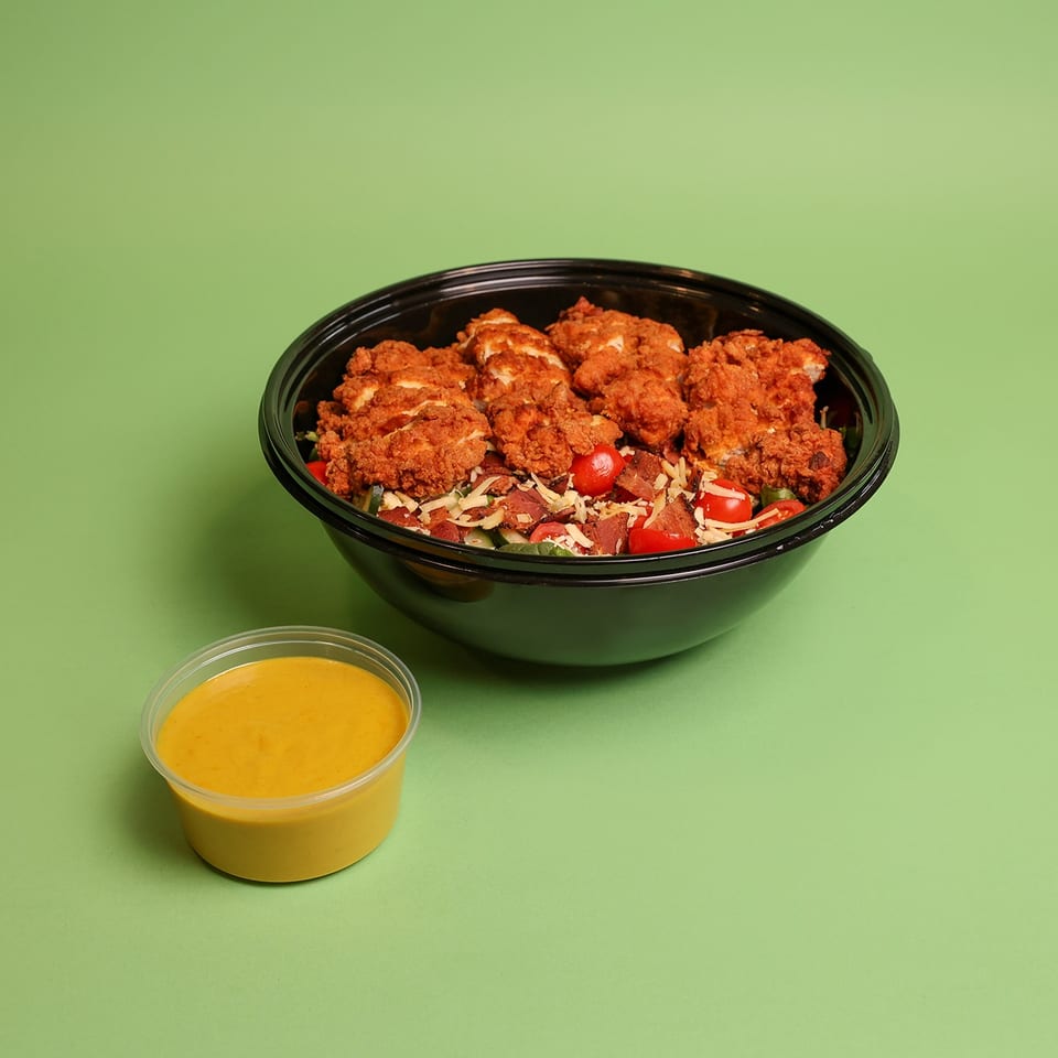 Crispy fried chicken salad with tomatoes, bacon, and shredded cheese in a black bowl, served with yellow dipping sauce.
