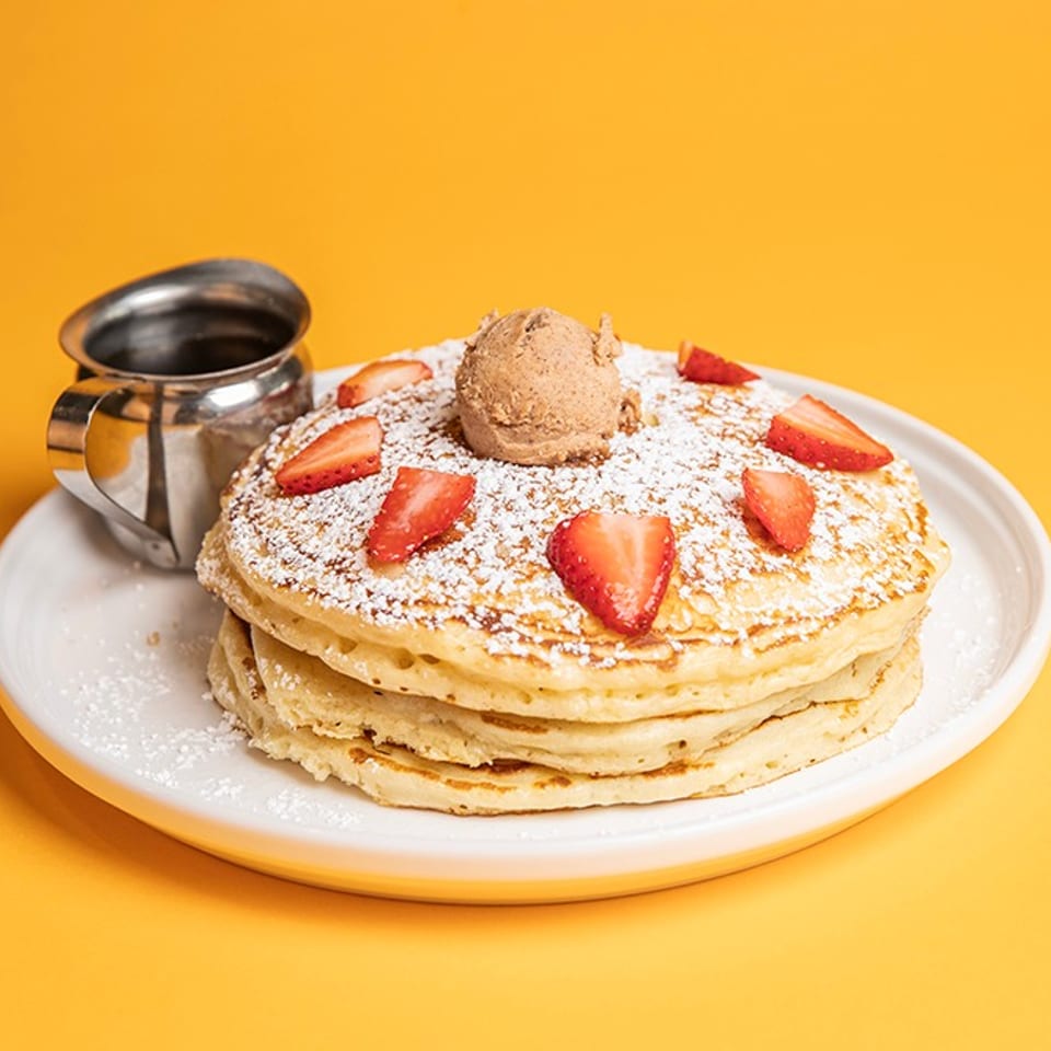 Stack of pancakes topped with strawberries, powdered sugar, and ice cream, served with syrup on a white plate.