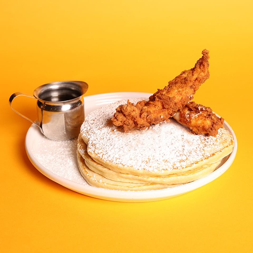 Stack of powdered-sugar pancakes topped with fried chicken tenders, served with a small metal syrup pitcher on a plate.