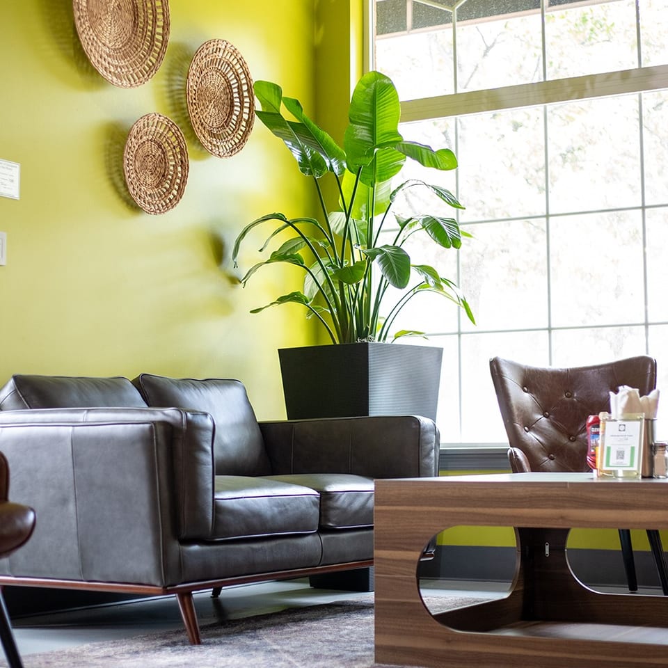 Bright restaurant lounge with leather sofas, a potted tropical plant, woven wall baskets, and a large arched window.