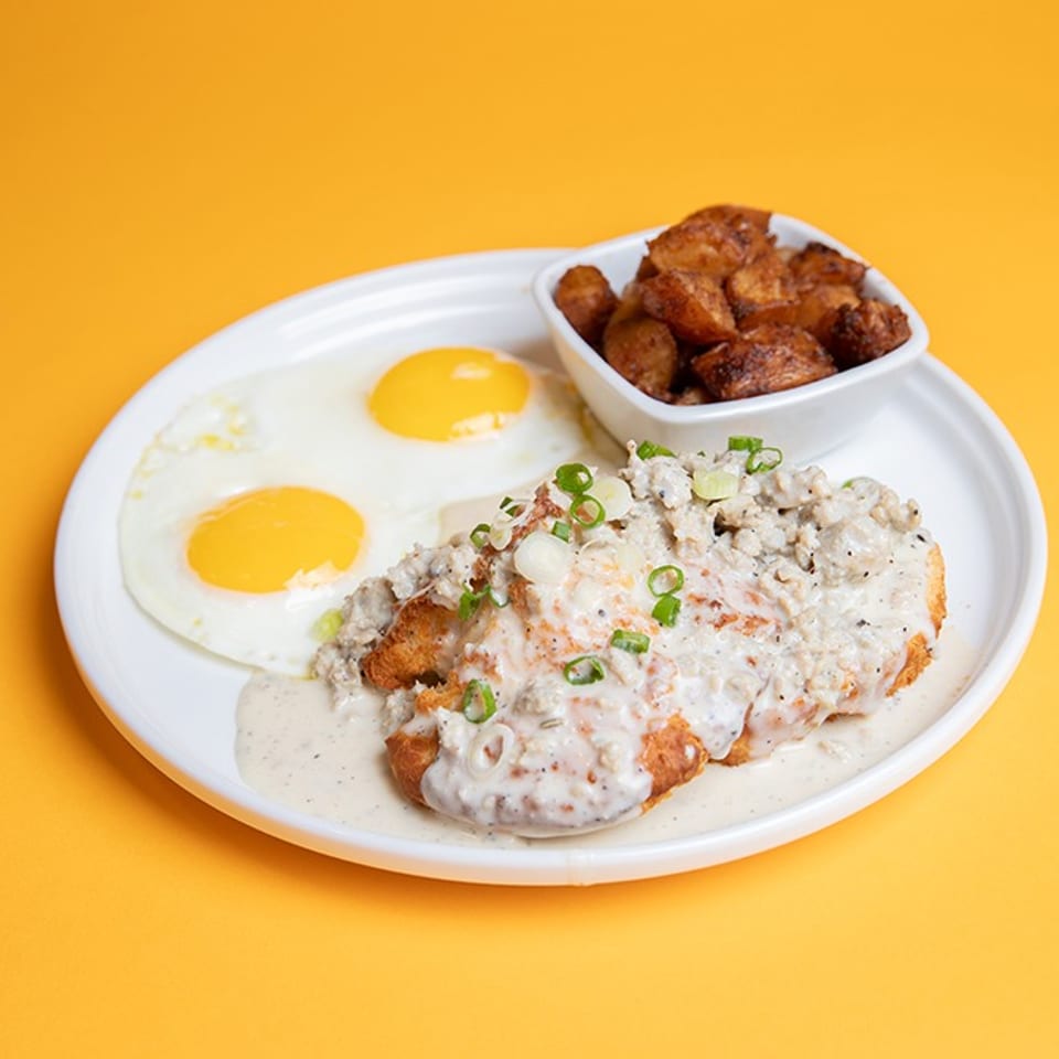 Plate of creamy country-fried steak topped with green onions, two fried eggs, and seasoned breakfast potatoes.