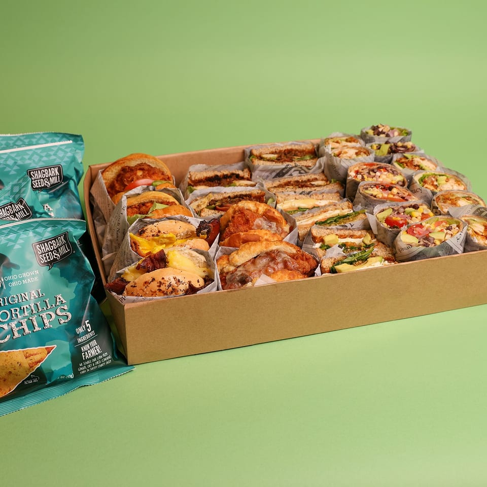 Box filled with assorted sandwiches and wraps beside teal Shagbark Seed & Mill tortilla chip bags on a green background.