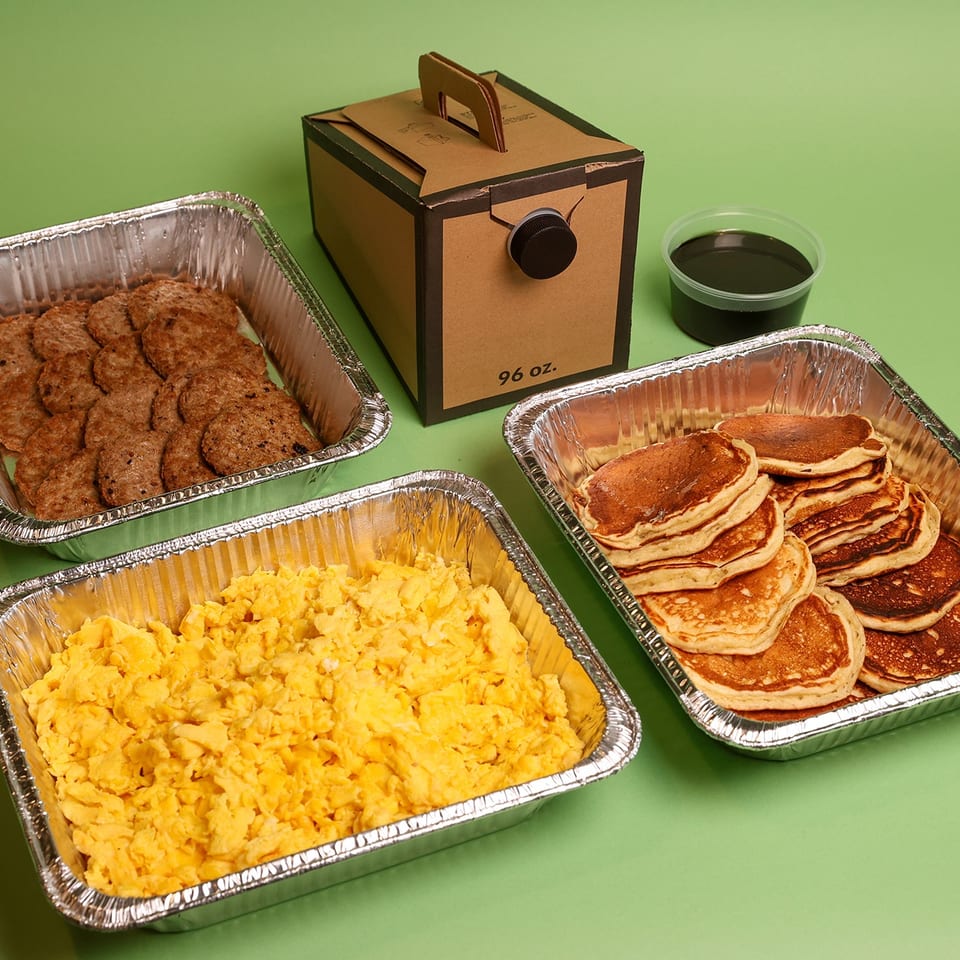 Catering spread with pancakes, breakfast sausage, scrambled eggs, syrup, and a labeled 96-ounce beverage container on a green background.