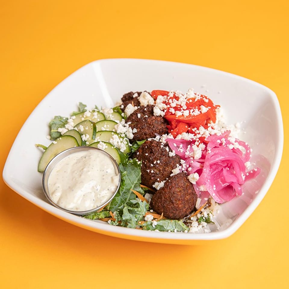 Mediterranean grain bowl with falafel, cucumber, tomatoes, pickled onions, feta, greens, and creamy dressing on an orange background.