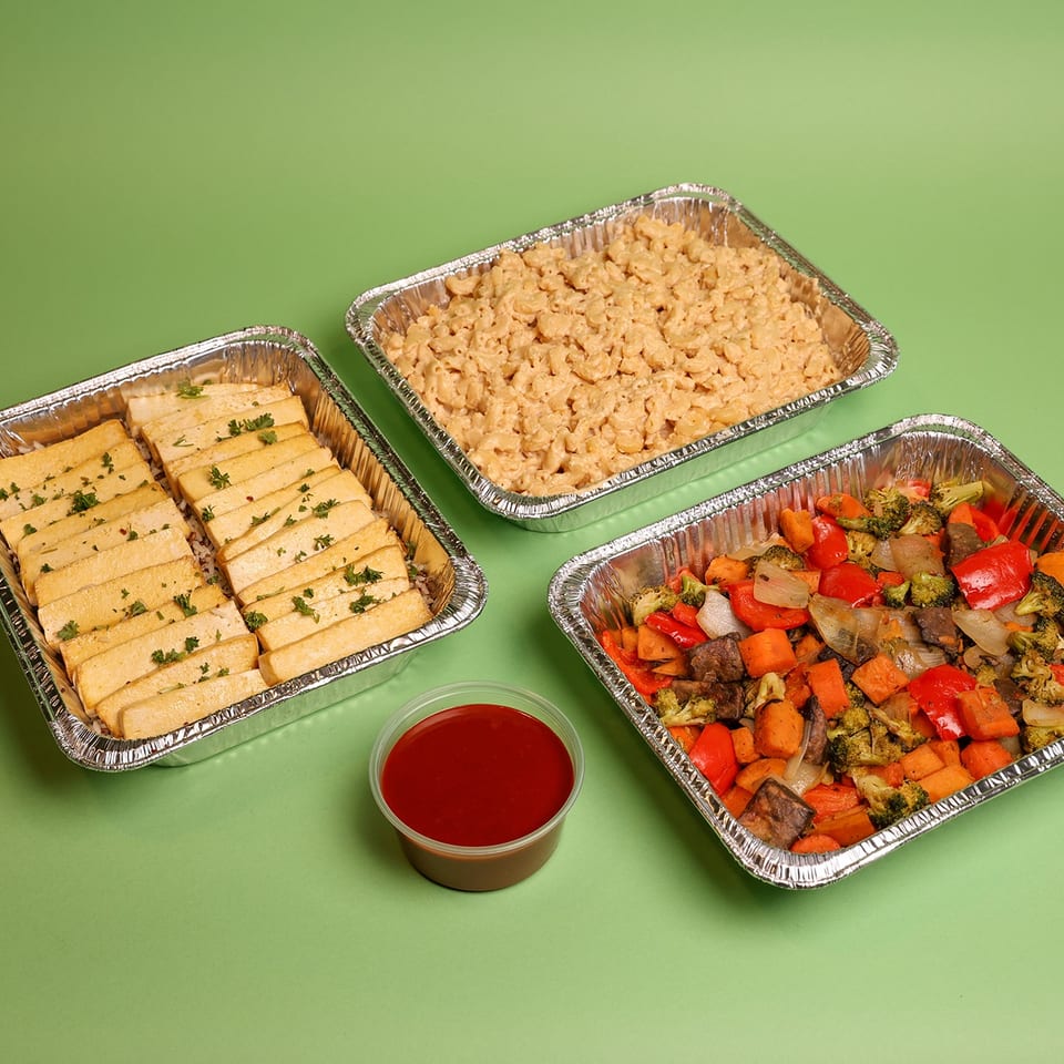 Three foil trays hold sliced herb-seasoned tofu, creamy macaroni, and roasted mixed vegetables with a cup of red sauce.