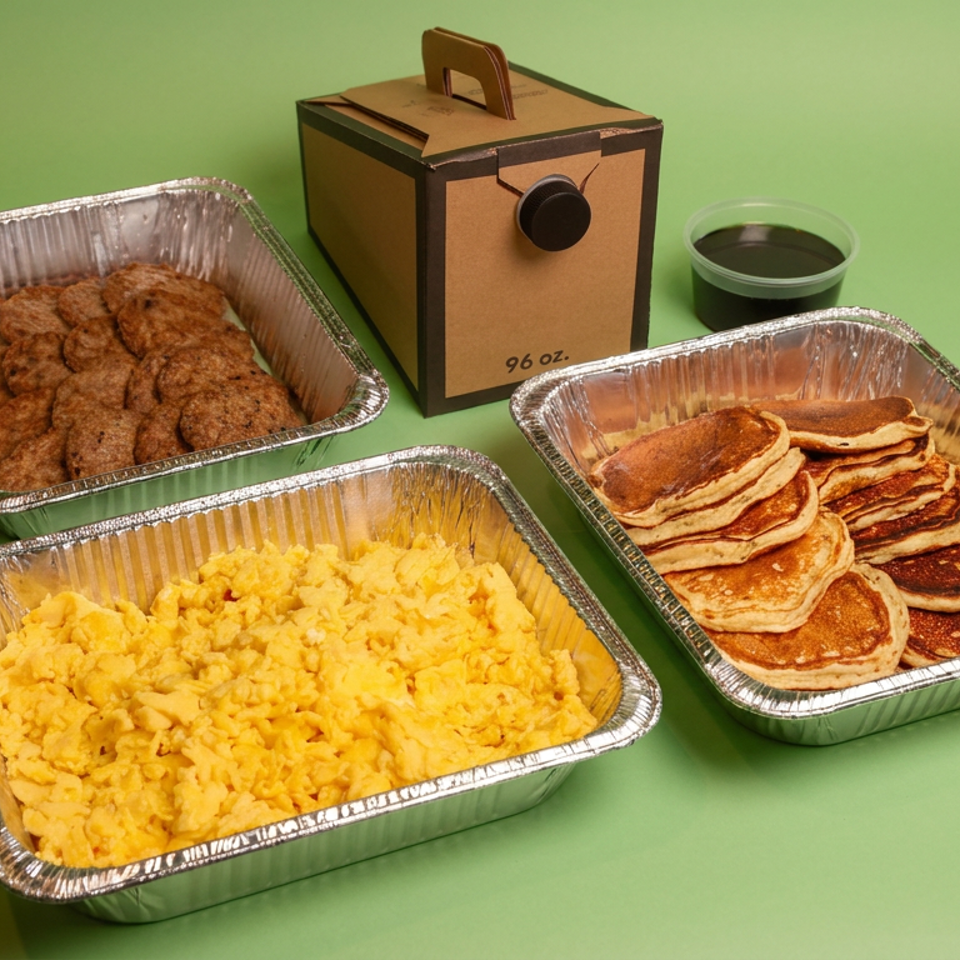 Catering spread with scrambled eggs, pancakes, fried chicken, syrup, and a wooden serving box arranged on a green background.