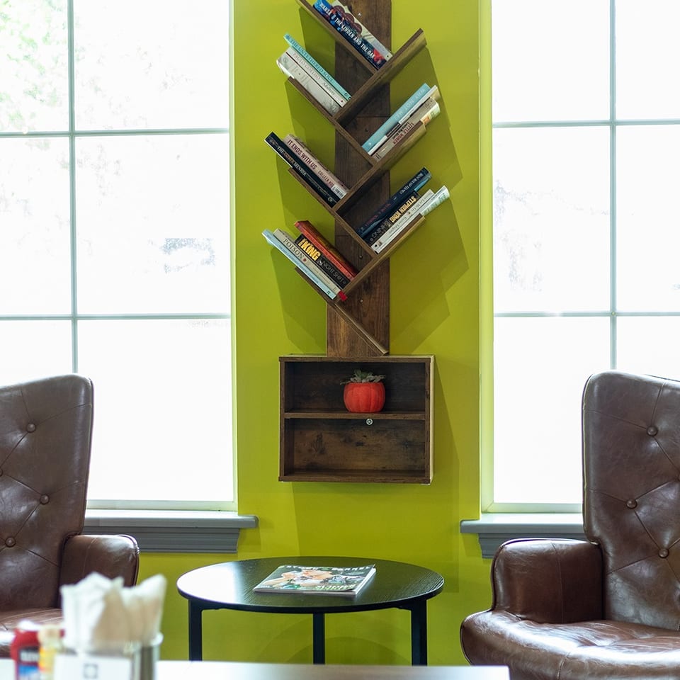 Restaurant seating area with two leather armchairs, a small round table, bright windows, and a wall-mounted bookshelf.