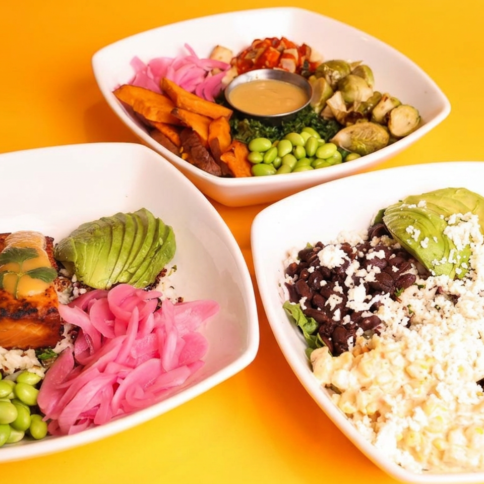 Three colorful grain bowls feature salmon, avocado, beans, edamame, pickled onions, roasted vegetables, sauces, and crumbly cheese on an orange background.