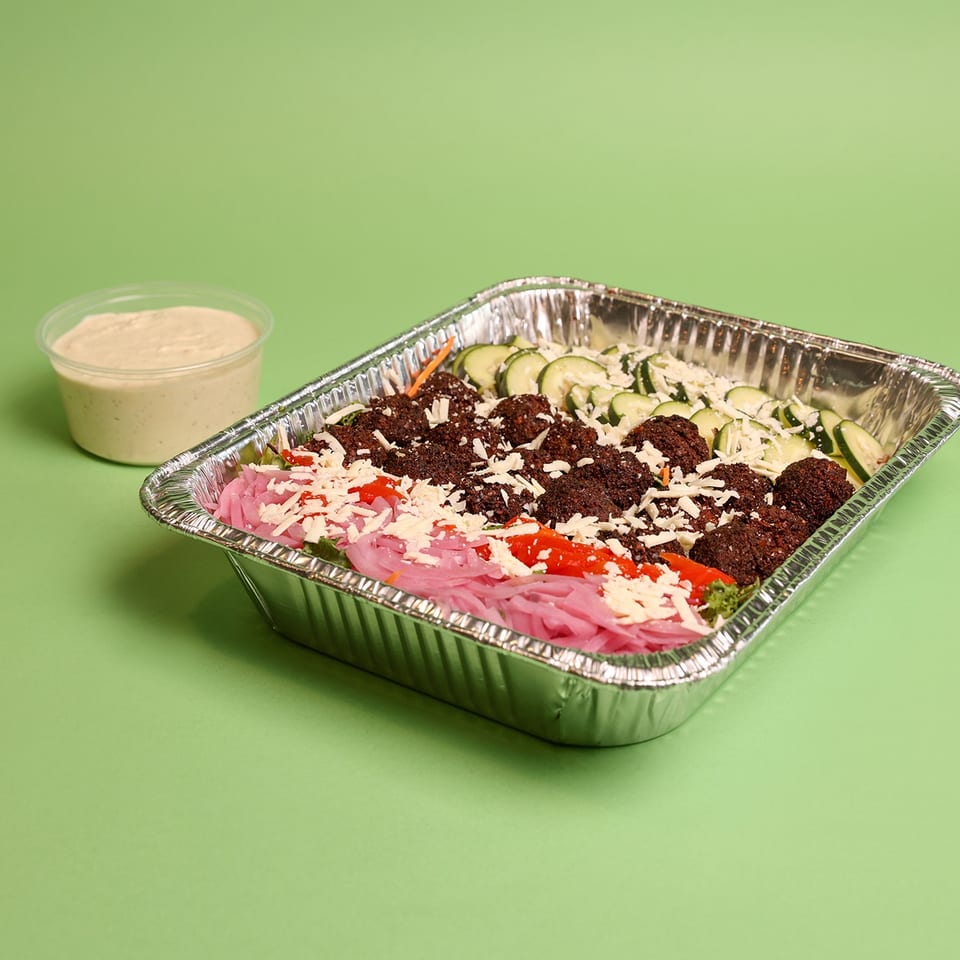 Aluminum tray of falafel, sliced cucumbers, pickled onions, peppers, and shredded cheese with tahini sauce on a green background.