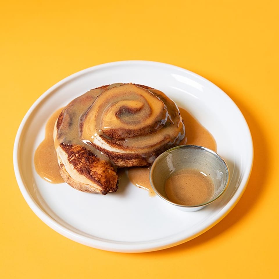 Cinnamon roll drizzled with caramel glaze on a white plate, accompanied by a small cup of sauce against a bright yellow background.